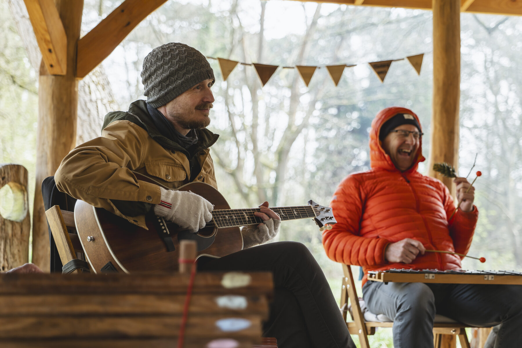 Two participants playing guitar and xylophone beneath a woodland shelter