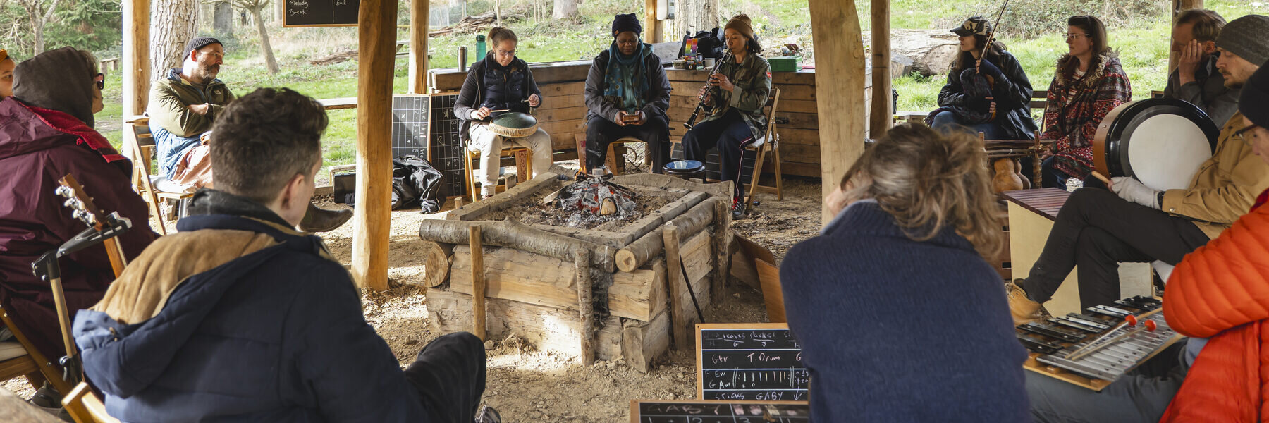 A group gathered around a fire beneath a woodland pavilion, making music together